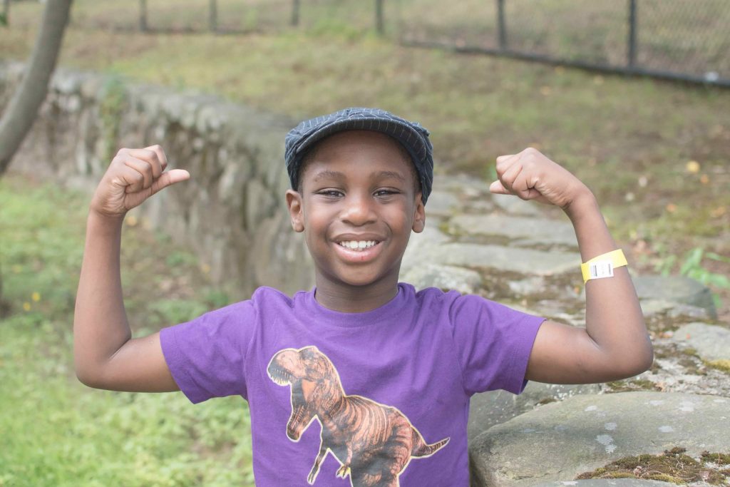 african american kid wearing a purple shirt with a t rex on the front flexing his muscles