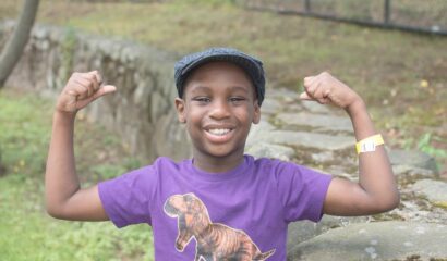 african american kid wearing a purple shirt with a t rex on the front flexing his muscles