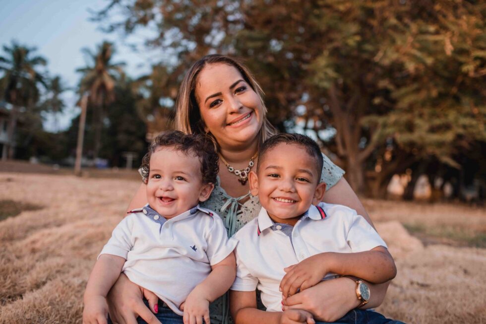 woman carrying two smiling young boys in her lap in the middle of a park