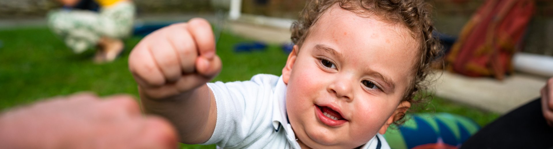 A smiling toddler reaches out toward a caregiver's hand